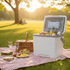 A 13L white cooler box on grass with drinks inside, surrounded by food and picnic items. It has a branding logo.