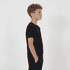 A black youth tee is displayed on a young person standing sideways with hands in pockets.