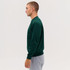 A green crew sweatshirt worn by a male model, paired with grey sweatpants, displayed from the side.