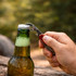 A stainless steel multi-function utensil opening a green drink bottle on a wooden surface in an outdoor setting.