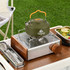 A green camping kettle with an orange handle sits on a silver stove beside a plate of food on a wooden table.