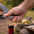 A silver multitool shaped like a fish, being used to open a bottle, with a nature setting in the background.