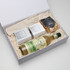 A white gift box containing a drink bottle, two small boxes, and a bag of pretzels, all in neutral tones.