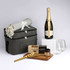 A grey hamper contains a bottle of sparkling wine, two glasses, a cheese board, a blanket, and gourmet snacks.