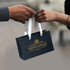 A small black paper bag with a white ribbon handle, featuring a logo in gold.