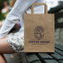A medium flat handle paper bag in brown with a logo, placed on a bench beside a person.