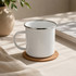 A white enamel mug rests on a round cork coaster, set on a light-coloured table with soft shadows and a plant in the background.