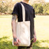 A beige tote bag with long handles is held by a person standing outdoors on grass. The bag has a logo.