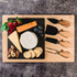 A bamboo cheese board featuring a variety of cheeses, fruits, nuts, and three wooden-handled knives.