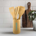 A bamboo cutlery set consisting of various utensils, displayed in a cylindrical holder, on a countertop with a plant and cutting board.