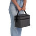 A grey cooler bag with a handle is being held by a person wearing light blue jeans. The bag features a front pocket and branding.