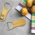 A drink bottle opener and peeler made from bamboo, featuring a metal bottle opener and surrounded by potatoes and a striped cloth.