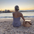 A wicker cooler basket in a light colour, positioned near a person on a beach at sunset. The scene features water and a cityscape.