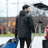 A man in a black rain jacket with a hood stands by a car, holding a sports bag and hockey stick. The jacket features a logo.