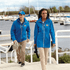 A blue rain jacket with a hood, featuring a logo, worn by a man and a woman by a marina.