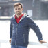 A man wearing a navy puffer jacket with a hood, standing by a waterfront. The jacket has a patterned interior and features a logo.