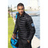A black puffer jacket with a zip front and a logo on the sleeve, worn by a model near water with a blue bag.