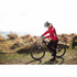 A woman in a red jacket and black cycling gear stands next to a mountain bike on a trail with a scenic background.