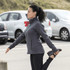 A woman stretches while wearing a grey polar fleece jacket, with parked cars in the background. The jacket features a logo.
