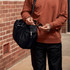 A black duffle bag is held by a person wearing an orange-brown sweatshirt, with sunglasses in their other hand.
