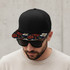 A black flat peak cap with a patterned floral brim, worn by a man in sunglasses.