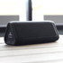 A black outdoor speaker with a triangular shape and a logo, resting on a wooden surface.