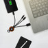 A round light-up charging cable in black and brown connected to a smartphone and laptop on a white surface.