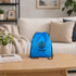 A blue drawstring bag sits on a wooden table in a cosy living room setting with a plant nearby. It features a logo.
