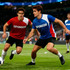 Two male models wearing reversible jerseys, one in red and the other in blue, both with a logo, on a sports field.