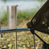 A drink bottle in stainless steel sits on a campsite table, with a logo on its side and a blurred background of nature.