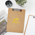 A wooden clipboard in a light brown colour with a silver metal clip. It features a logo and is placed on a desk with greenery.