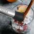 A long stainless steel cocktail muddler with a black silicone tip sits above a glass filled with ice and apple slices.