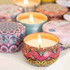 A collection of colourful tin candles with intricate designs and lit wicks, arranged on a white surface.