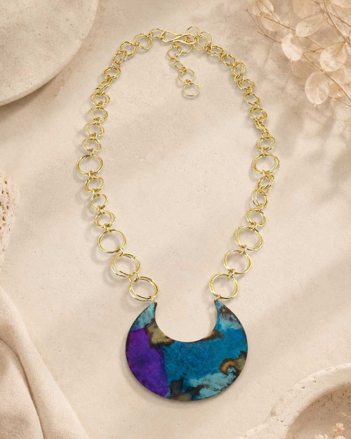 Hand-patinated brass crescent pendant necklace arranged flat on a textured beige surface with dried botanical props nearby