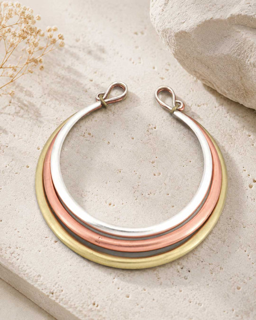 Tri-Tone Brass Collar Necklace laid flat on a textured stone surface beside dried flowers, showing silver, copper, and brass rings