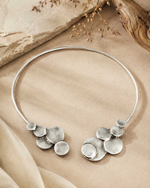 Pewter collar necklace with disc clusters at both open ends, laid flat on a sandy stone surface with dried botanicals