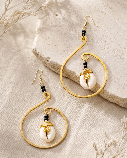 Cowrie shell and hammered brass hoop earrings laid on a stone surface with dried botanicals, angled view showing wire-wrapped shells