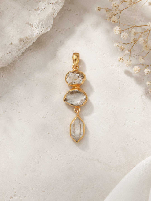 Triple Herkimer Quartz gold-plated pendant on a stone surface with dried flowers, showing three stacked crystals in gold bezels