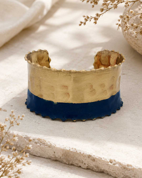 Hand-hammered brass cuff bracelet with indigo painted lower band and scalloped edges, front view on stone surface with dried botanicals