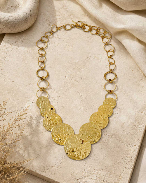 Hammered brass cascading disc necklace laid flat on a stone surface with dried grasses, styled flat lay view