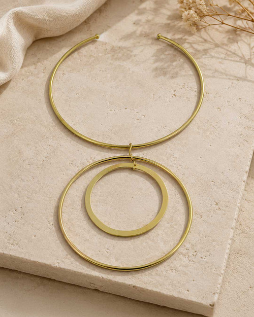 Brass collar necklace with double ring pendants laid flat on a stone surface with linen and dried florals