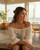 Hand-forged brass disc blue patina necklace worn by a woman in a white off-shoulder top, seated near an ocean-view window, looking sideways