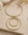 Brass collar necklace with double ring pendants laid flat on a stone surface with linen and dried florals