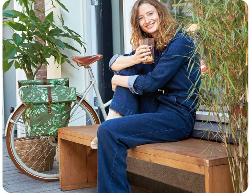Smiling woman in denim sitting on a wooden bench, holding a glass, with a bicycle equipped with a green patterned bag in the background.