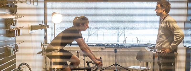 A bike fitting session with a cyclist on an indoor trainer and an instructor observing in a well-lit room.
