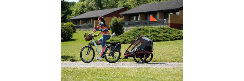 Woman riding a bike with a child trailer attached, on a grassy path near residential houses.