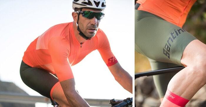 A cyclist wearing a bright orange cycling jersey and olive-green shorts with 'Santini' branding, paired with a white helmet and sunglasses, riding outdoors in natural light