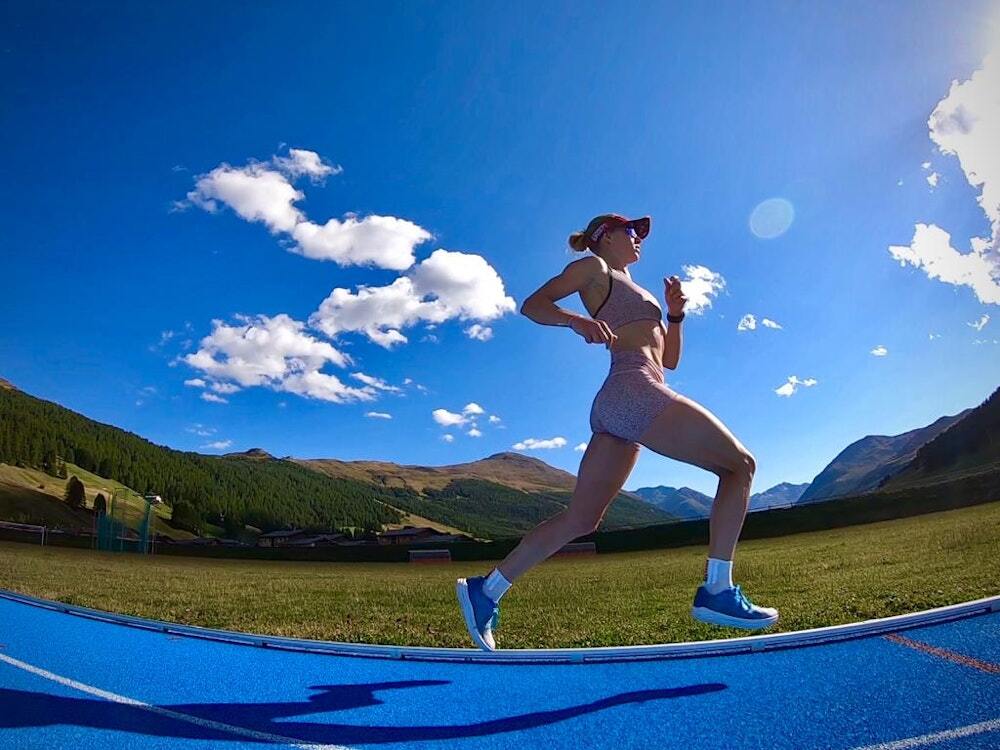 A female runner in athletic gear mid-stride on a bright blue running track, surrounded by mountains under a clear blue sky