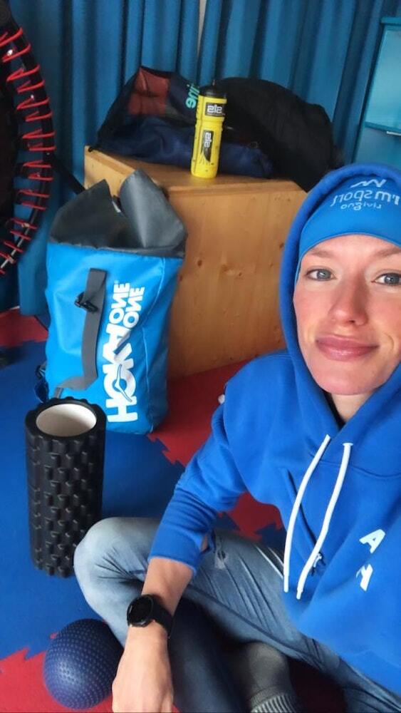 A woman in a blue hoodie sitting on the floor of a gym, surrounded by fitness equipment, including a foam roller, exercise balls, and a yellow SiS water bottle