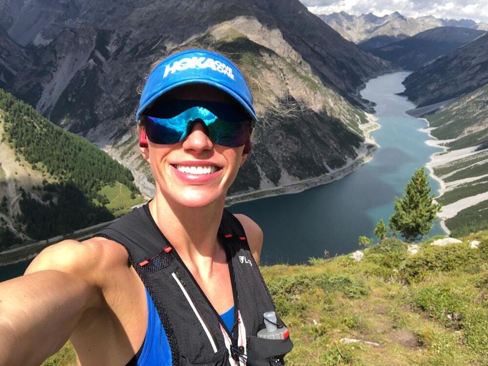 A woman in a cap and sunglasses smiling for a selfie on a mountain trail